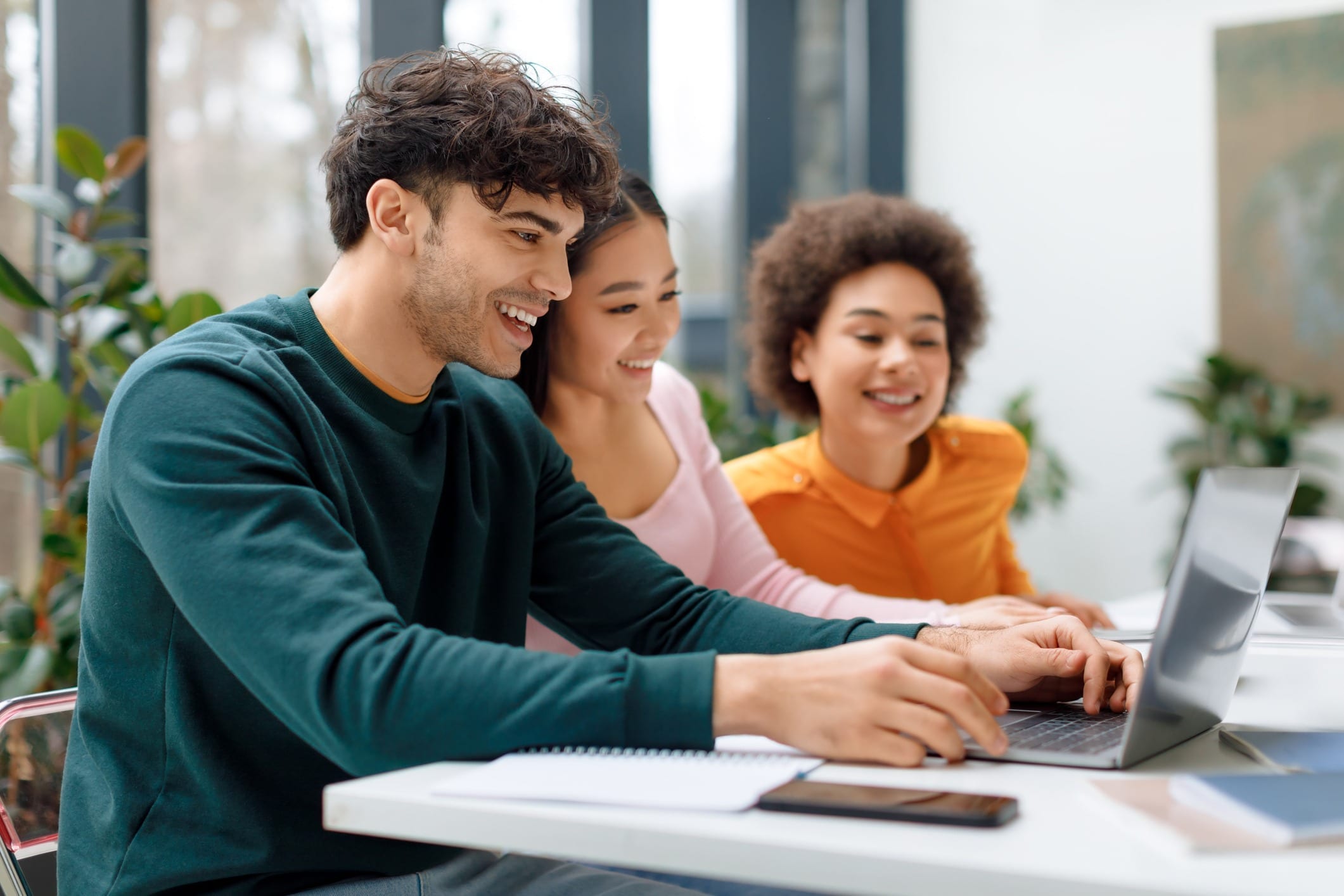Three students smiling at a laptop together beside a sunlit window