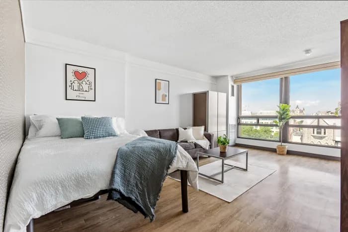 Bright and modern studio apartment with a cozy bed, stylish couch, and large window showcasing a stunning city view.